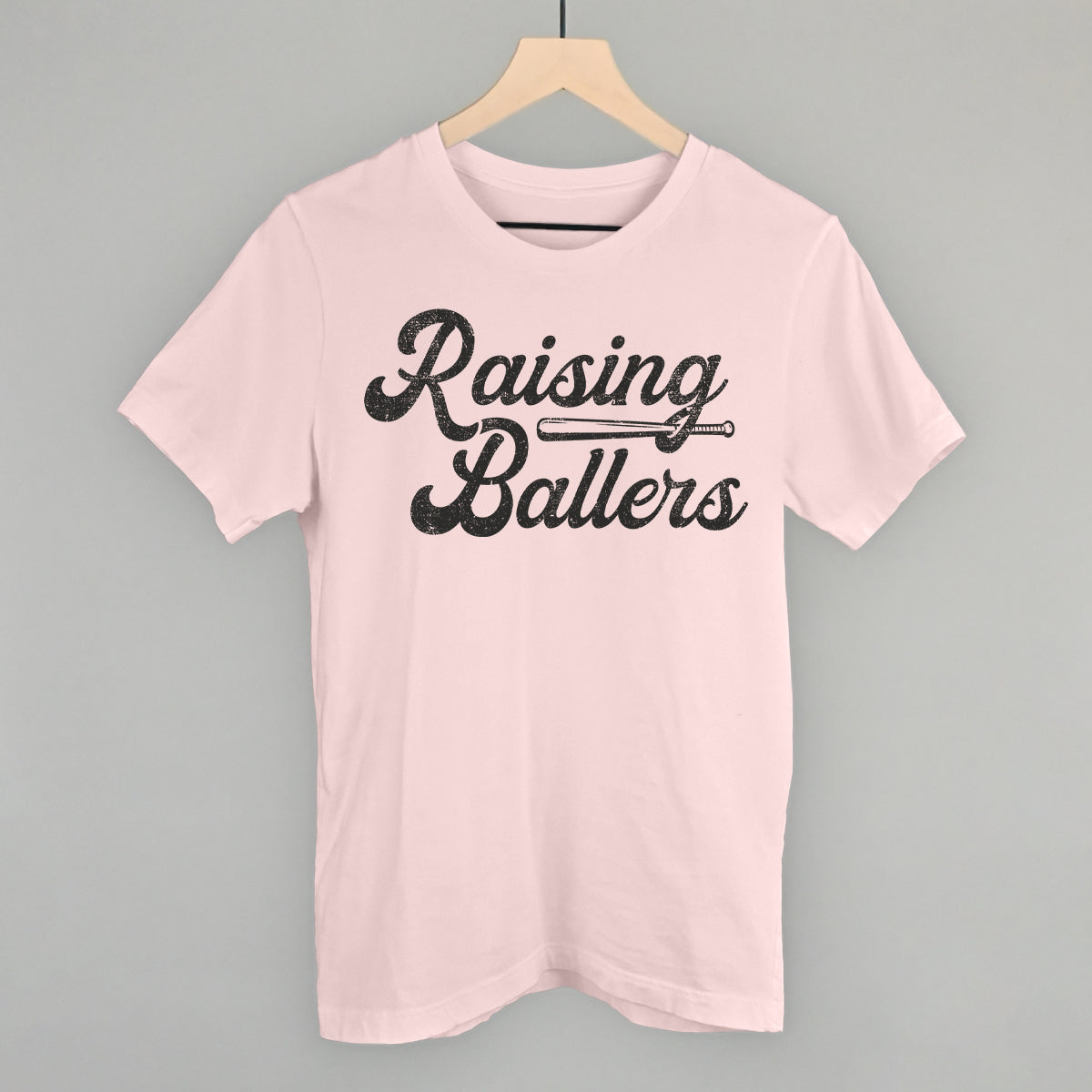 Pink t-shirt with 'Raising Ballers' text on a hanger against a gray background