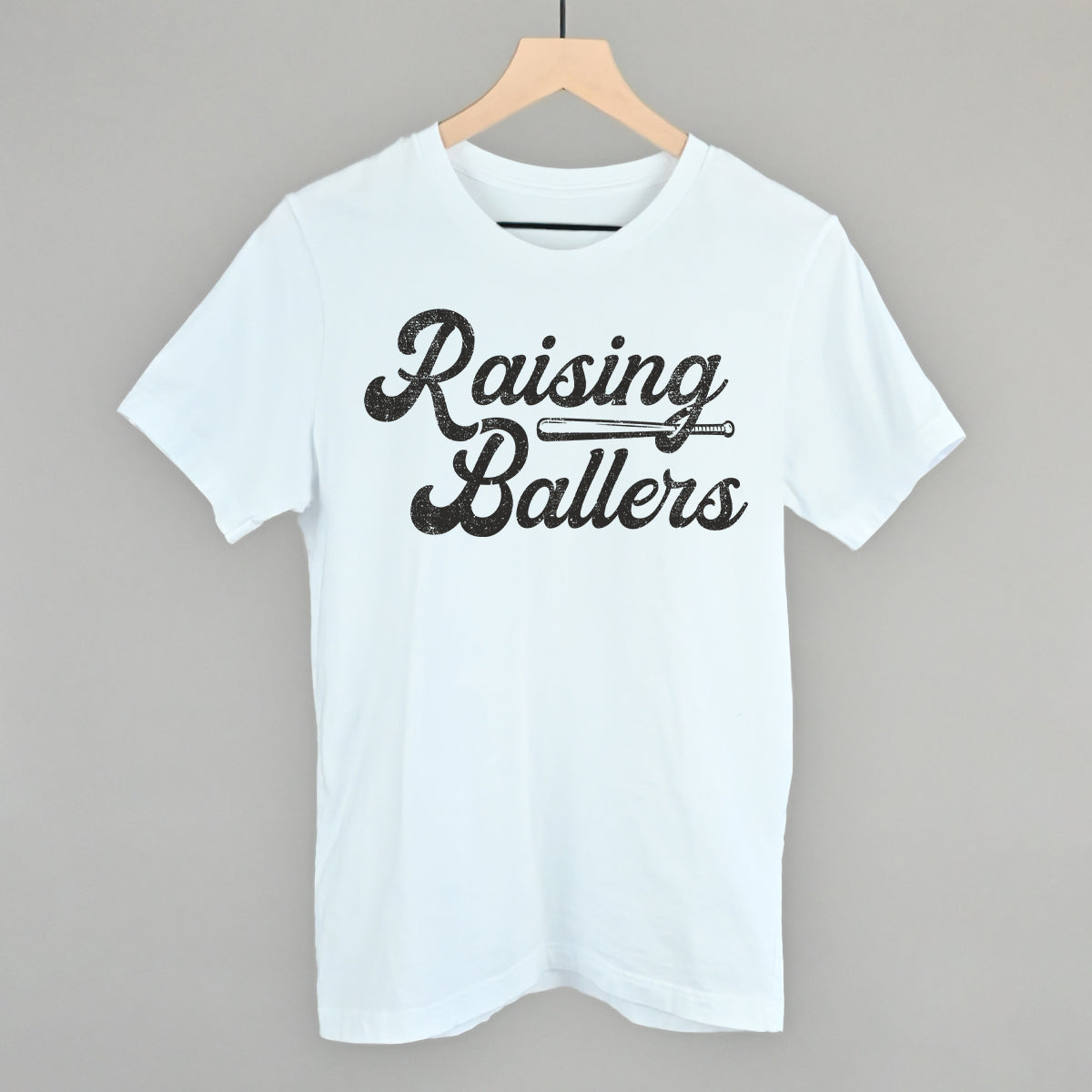 White t-shirt with 'Raising Ballers' text on a hanger against a gray background