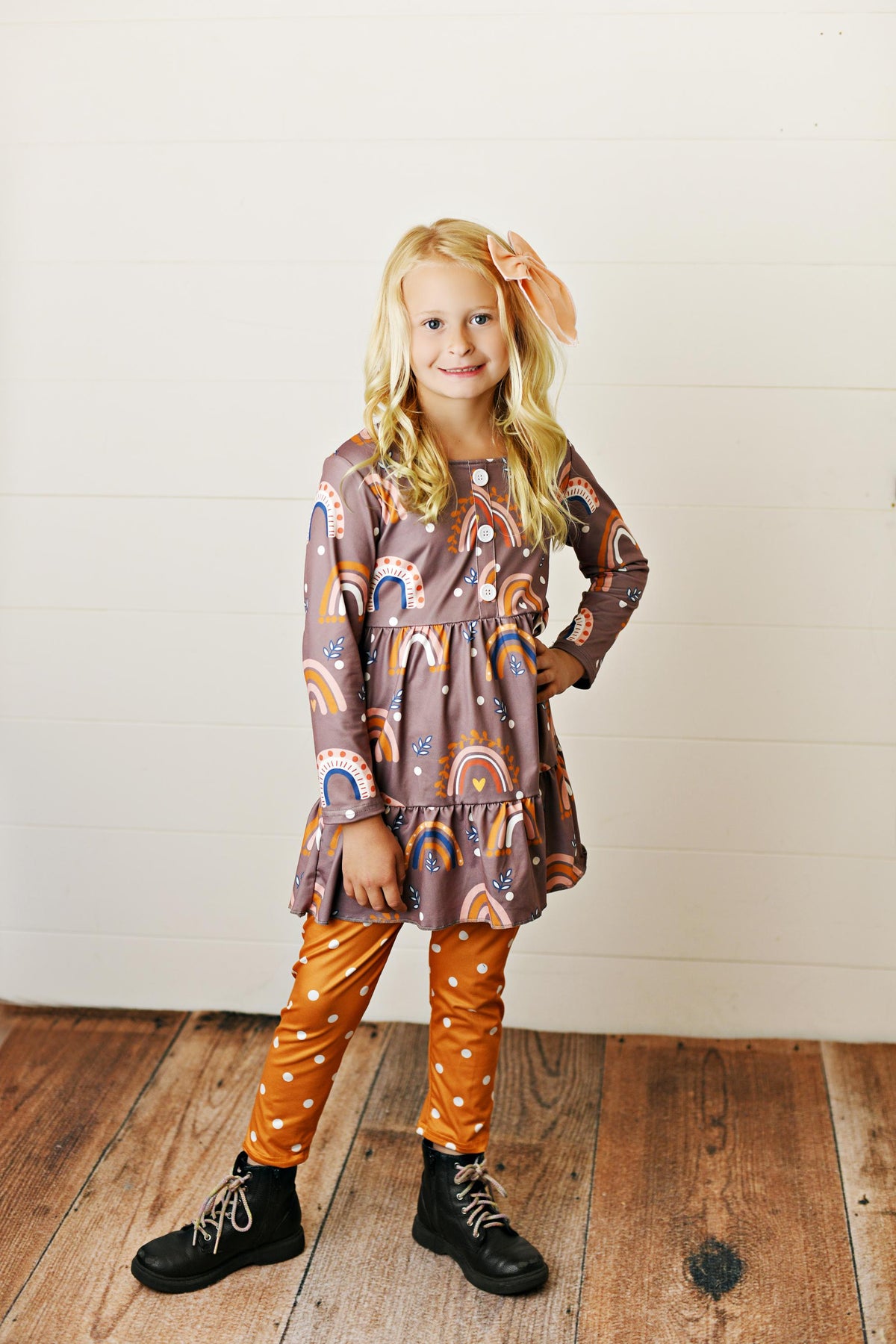 A young girl with blonde hair wearing a brown tiered long-sleeve dress with a colorful rainbow pattern, orange polka-dot pants, and black boots, standing on a wooden floor against a white paneled wall.