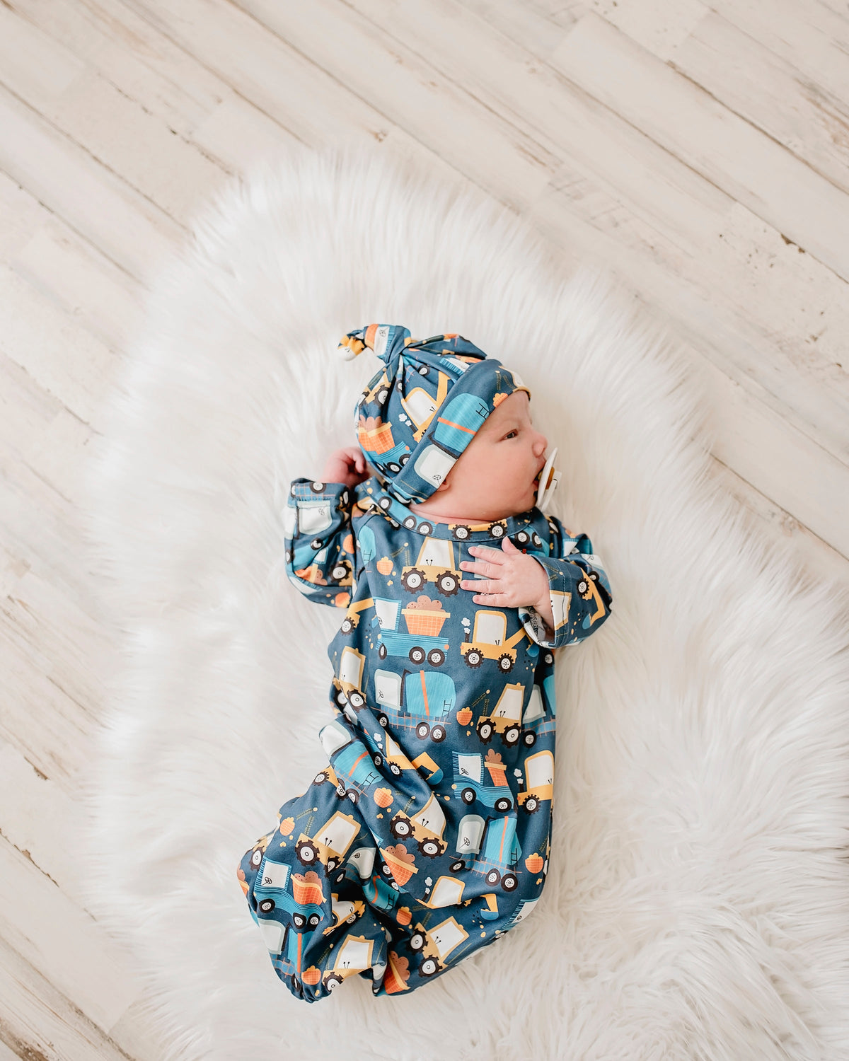 A newborn baby wearing a blue long-sleeve romper and matching knotted bonnet with a truck print, lying on a white faux fur rug over a light wood floor.