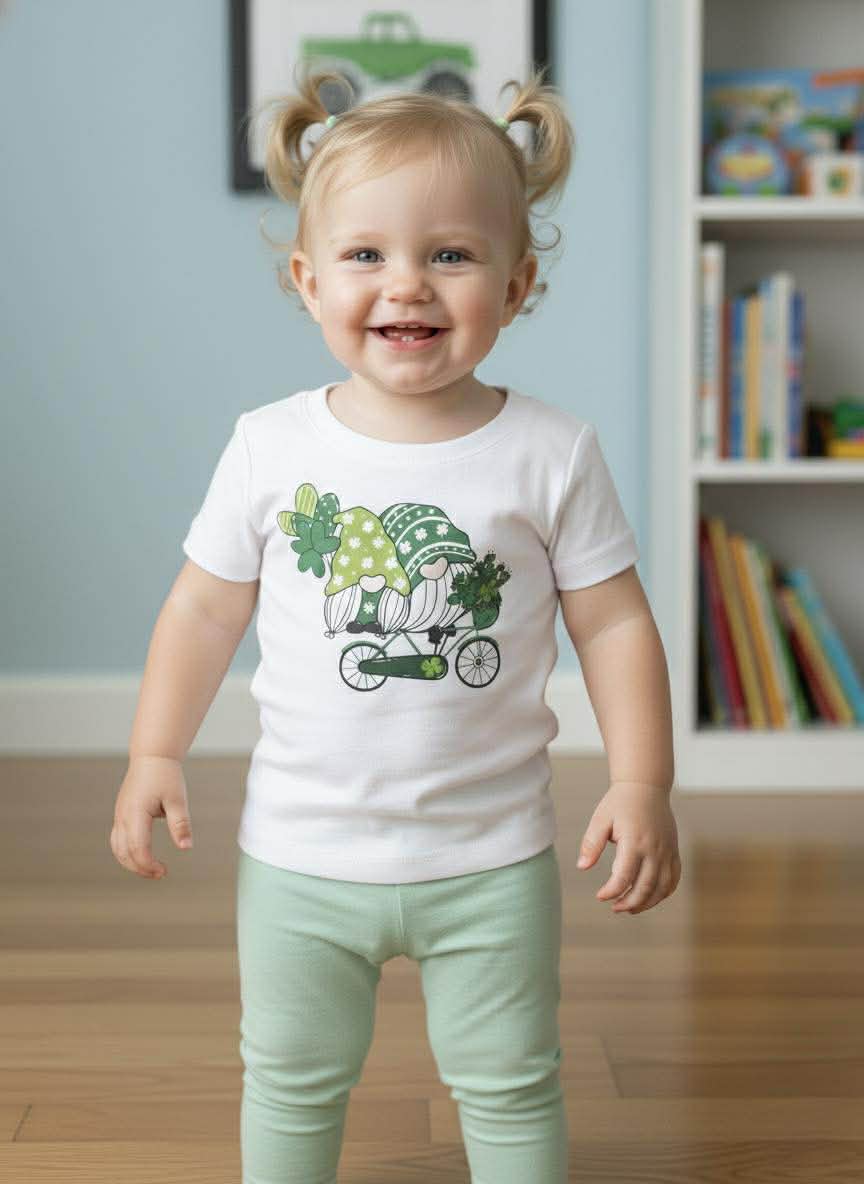 Child wearing a white t-shirt with a green graphic design, standing in a room with a bookshelf in the background.
