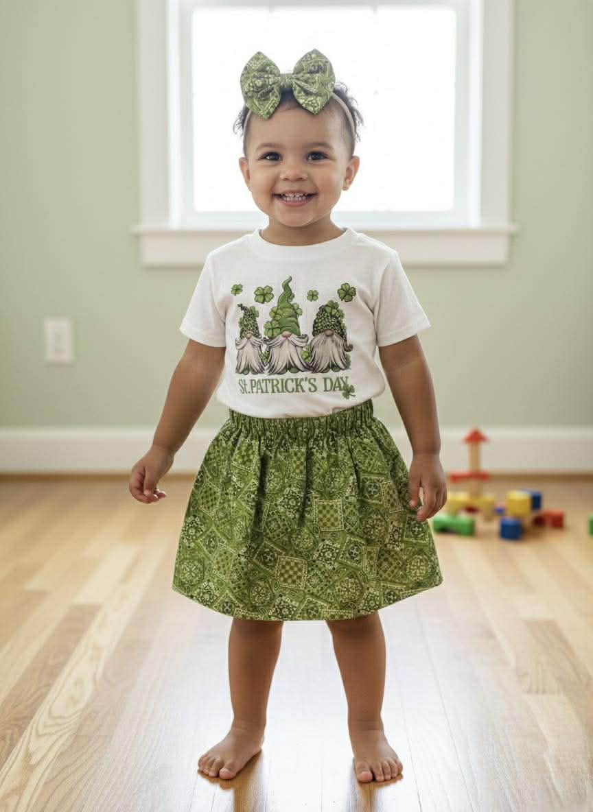 Child wearing a St. Patrick's Day shirt and green skirt in a room with toys on the floor.
